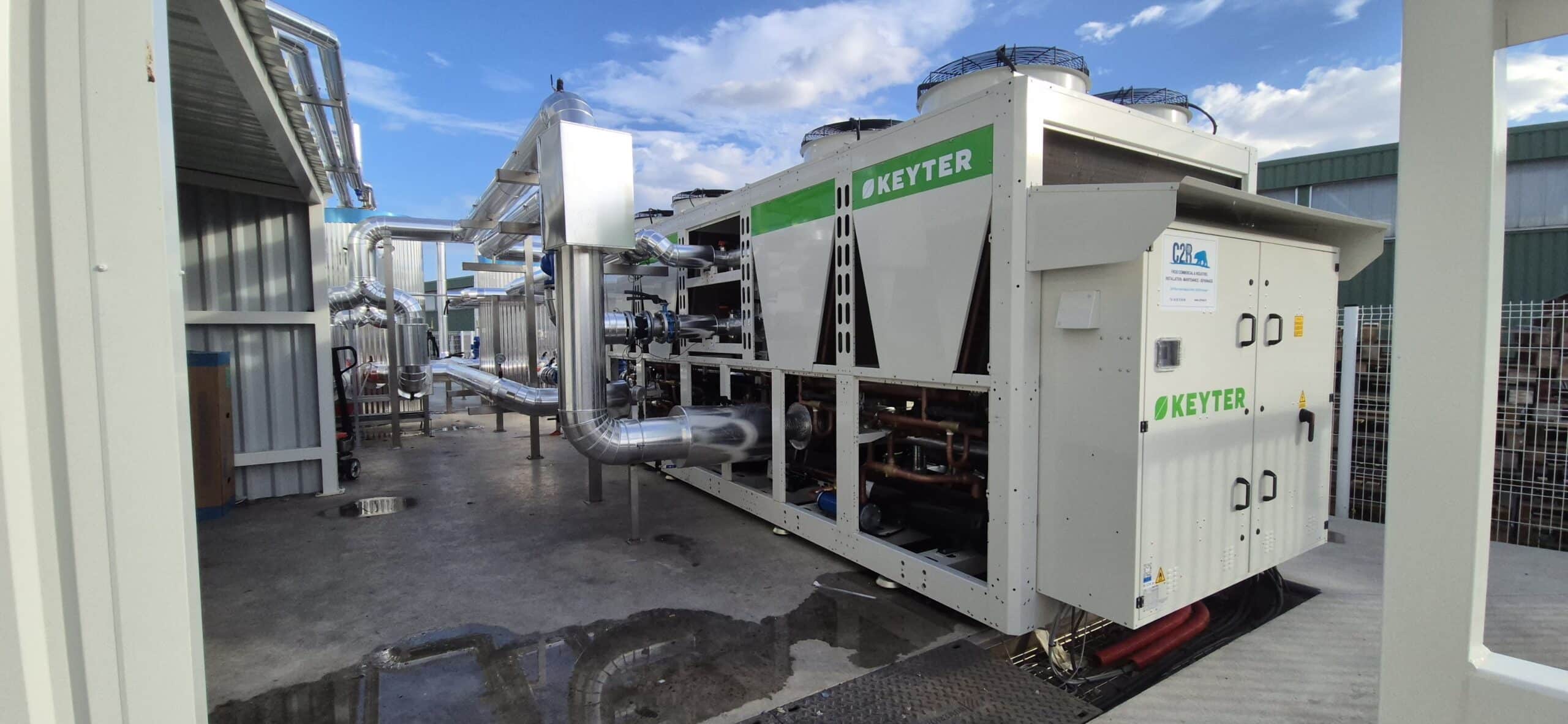 Chiller industriel KEYTER blanc et vert avec logo C2R, raccordé à une tuyauterie isolée en extérieur. Installation de froid commercial sous ciel nuageux.